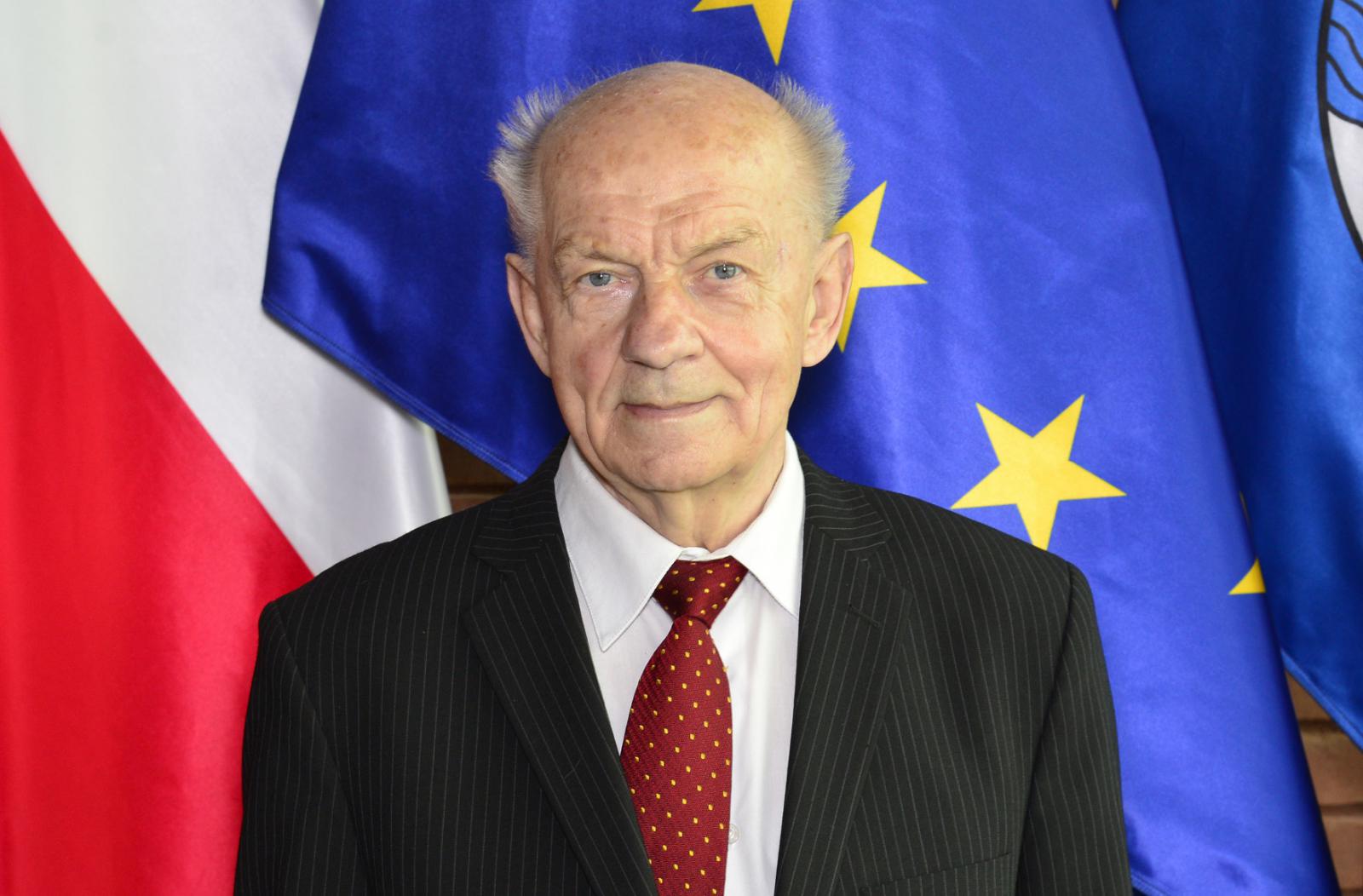 Zdjęcie portretowe mężczyzny – krótkie jasne włosy, ubrany jest w białą koszulę, czarną marynarkę i bordowy krawat. W tle widoczne fragmenty flag.