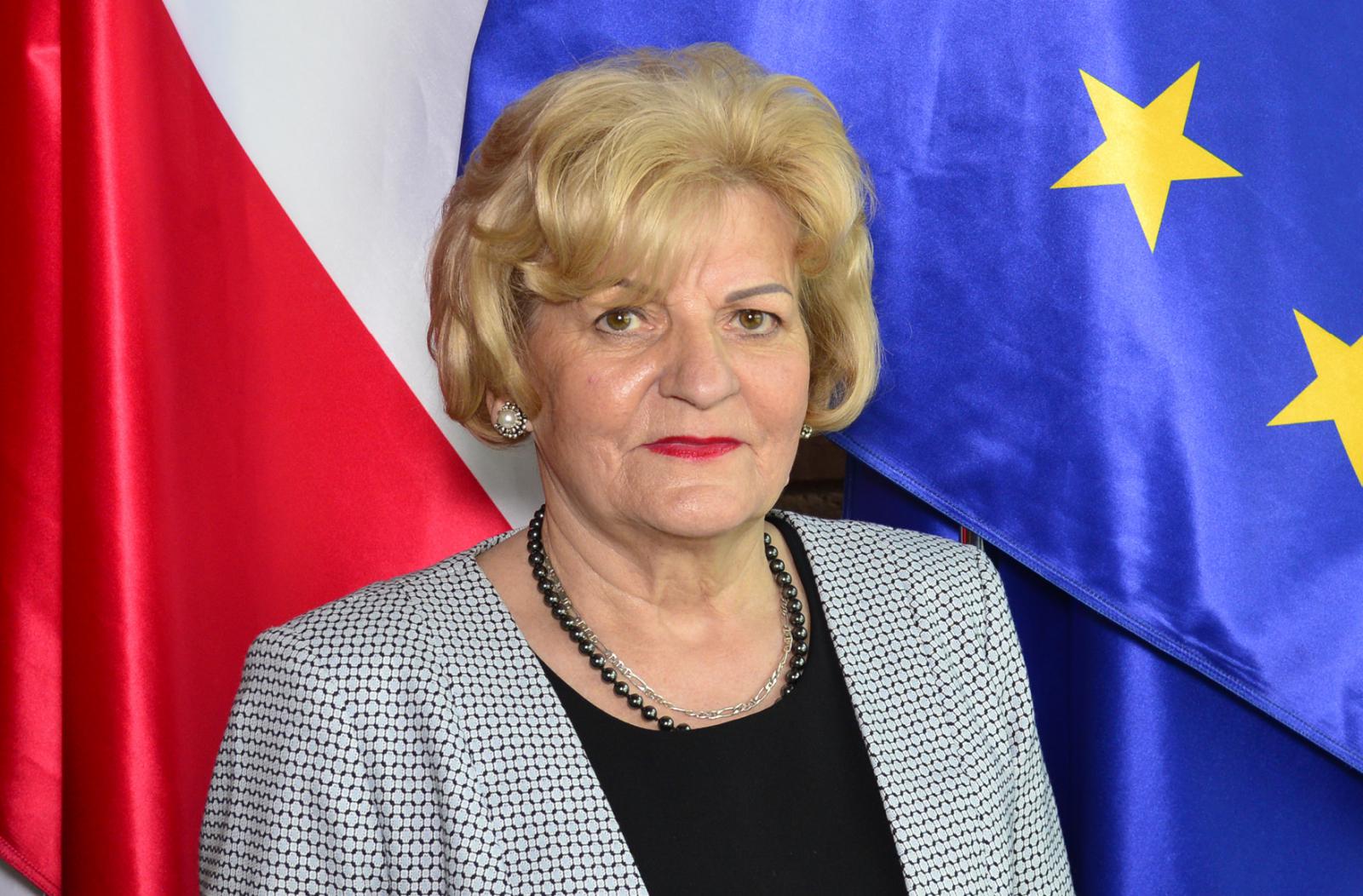Zdjęcie portretowe kobiety – krótkie jasne włosy, ubrana jest w szarą, wzorzystą marynarkę i czarną bluzkę. W tle widoczne fragmenty flag.