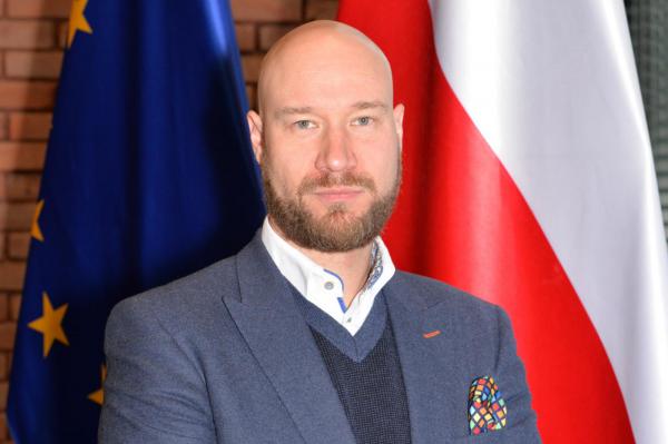 Zdjęcie portretowe mężczyzny – ciemne wąsy i broda, ubrany jest w niebieską marynarkę, ciemny sweter i białą koszulę.