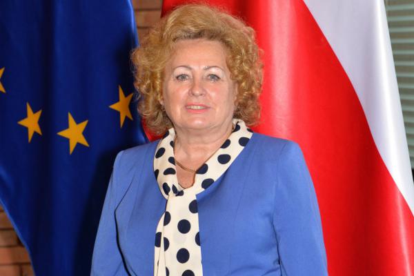 Zdjęcie portretowe kobiety – jasne kręcone włosy, ubrana jest w niebieską marynarkę, na szyi biały szal w czarne kropki. 