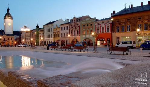 Widok na rynek miejski, z zabytkową zabudową, po prawej stronie szereg kamieniczek, na lewo fragment budowli sakralnej z wysoką wieżą. W centralnej części znajduje się duża fontanna.