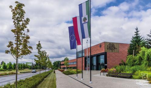 Duży, nowoczesny budynek użyteczności publicznej  ukazany z dużej perspektywy narożnika, ceglane ściany, duże okna i oszklenia, przed budynkiem drzewa oraz horągiew Konstancina-Jeziorny, flagi Polski i Unii Europejskiej na masztach 
