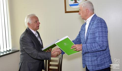 Dwaj mężczyźni w garniturach stoją za biurkiem, w lewych dłoniach trzymają zielone teczki na dokumenty, prawe dłonie są w uścisku. Za nimi widoczny jest fragment krzesła oraz wiszący na ściennie herb gminy.
