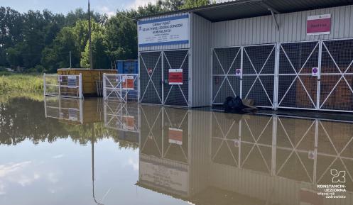 Na pierwszy planie zalany plac punktu selektywnej zbiórki odpadów komunalnych. W oddali, po lewej stronie, fragment altan śmietnikowych z kontenerami na odpady.