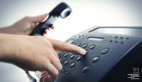Stacjonarny aparata telefoniczny. Widoczne są dłonie dorosłej osoby, która prawą ręką naciska przyciski z cyframi, a w lewej trzyma słuchawkę. 