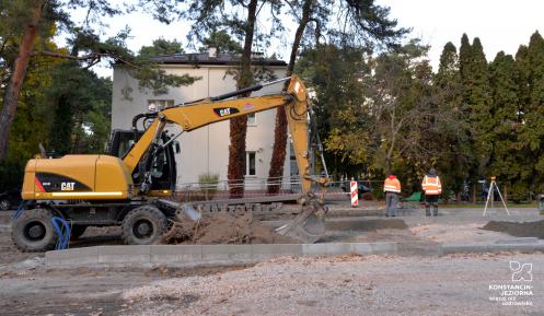 Remont drogi. Widać rozkopaną ziemię, obok stoi żółta koparka. Przy koparce stoi dwóch robotników, którzy ubrani są w pomarańczowe kurtki z odblaskami.