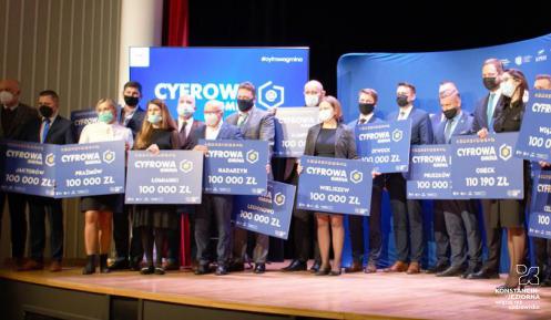 Na scenie stoi grupa osób (kobiety i mężczyźni) w maseczkach trzymających w rękach symboliczne czeki, na których jest tekst: Cyfrowa Gmina, nazwa gminy i 100 tys. zł.