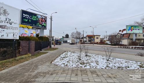 Ulica dwukierunkowa w Konstancinie-Jeziornie. Po ulicy jadą samochody, po chodnikach idą piesi. W dali widać budynki oraz duże banery reklamowe. 