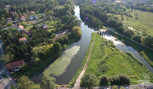 Zdjęcie miasta z lotu ptaka. W dolnej części widać ulicę i jadące po niej samochody. Obok ulicy płynie rzeka. W dali widoczne są zabudowania jednorodzinne.