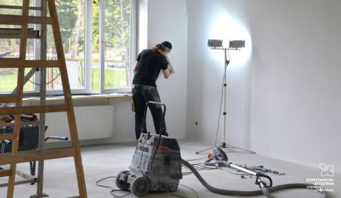 Pomieszczenie w remoncie, na pierwszym tle po lewej drabina, w środku odkurzacz. W głębi pracownik szlifujący ścianę. 