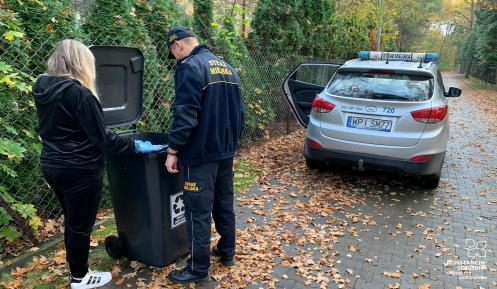 Na zdjęciu kobieta i mężczyna w policyjnym mundurze, zagladają do kosza z odpadami komunalnymi