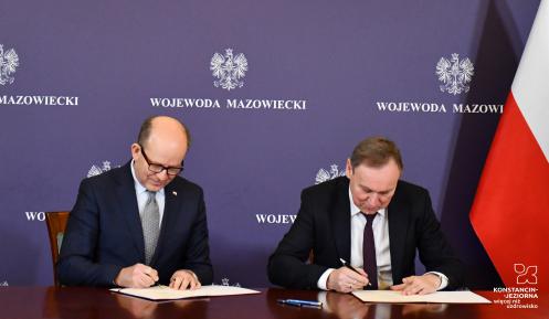  Dwaj mężczyźni, ubrani w garnitury, siedzą przy stole i podpisują dokumenty. 