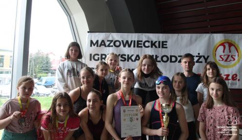 W pomieszczeniu obok dużego okna pozuje do zdjęcia 13 dziewczynek i jeden mężczyzna. Dziewczynki na szyjach mają medale, a jedna z nich złoty puchar w ręce. Za nimi wisi bilbord z napisem: Mazowieckie Igrzyska Młodzieży.. 