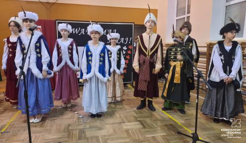 W sali stoi grupa dzieci w strojach ludowych i szlacheckich. Po prawej stronie na ścianie są drabinki gimnastyczne, przed nimi stojaki na mikrofony.