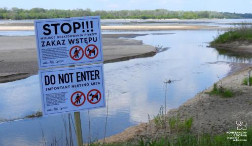Tabliczka z napisem: Stop. Zakaz wstępu. Za nią widać rzekę i plażę przy jej brzegach.