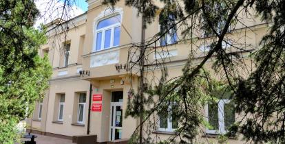 dwupiętrowy budynek willi z beżową elewacją i białymi oknami. Wokół gęste zadrzewienia częściowo zasłaniające budynek