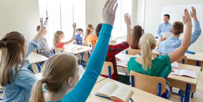 grupa młodzieży siedząca w klasie przy ławkach. Uczniowie mają uniesioną rękę do odpowiedzi. Przed nimi stoi uśmiechnięty nauczyciel.