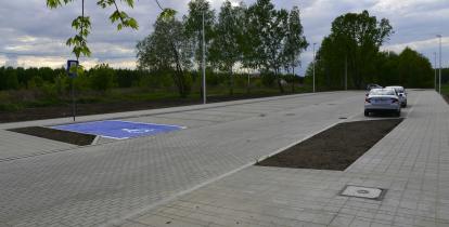 duży parking utwardzony kostką brukową oraz wyznaczonym miejscem dla osoby niepełnosprawnej. Po prawej ptronie wzdłuż chodnika są równolegle zaparkowane srebrne samochody. W tle gęste zadrzewienia. 