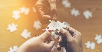 Trzy dłonie trzymające białe puzzle połączone ze sobą. W tle rozmazane wysypane puzzle. 
