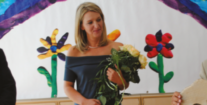 Kobieta – blond włosy do ramion, ubrana jest w niebieską sukienkę. W rękach trzyma bukiet białych róż. 