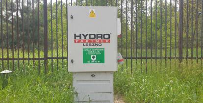 Metalowa skrzynka z napisem: Hydro Partner Leszno, Zakład Gospodarki Komunalnej. Wokół rośnie zielona trawa. Za nią metalowy płot.