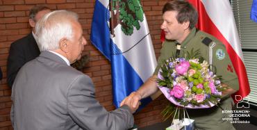 Dwie osoby w sali posiedzeń. Mężczyzna w szarym garniturze podaje rękę w geście gratulacji mężczyźnie w mundurze harcerskim.