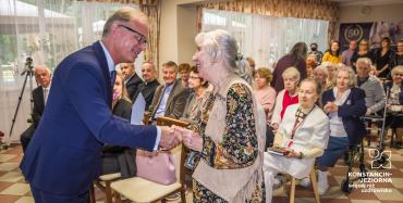 Sala. Mężczyzna w średnim wieku ubrany w garnitur oraz seniorka stoją naprzeciwko siebie, ściskają sobie prawe dłonie. Za nimi na krzesłach siedzi około dwadzieścia osób.