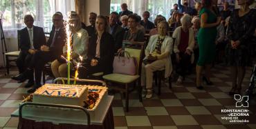 Sala. Około dwadzieścia osób siedzi na krzesłach. Z tyłu siedzą seniorzy. Z przodu siedzą dwie kobiety oraz dwóch mężczyzn w średnim wieku. Przed nimi na stoliku stoi duży biały tort z napisem Polska Akademia Nauk.