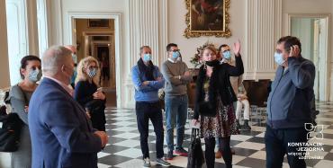 Muzeum. Dziesięć osób stoi w sali, twarze są zakryte maskami ochronnymi. Na ścianach wiszą obrazy z wizerunkami postaci historycznych oprawione w złote ramy.