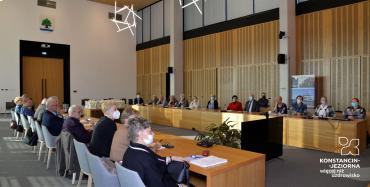 Sala posiedzeń. Przy dwóch drewnianych stołach ustawionych naprzeciwko siebie siedzi około trzydzieści osób, seniorzy i seniorki. 