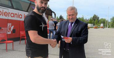dwie osoby, ściskają sobie dłoń, jedna z osób trzyma w ręku legitymację, w tle krwiobus