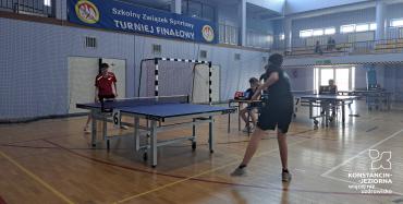 Na fali sportowej stół do tenisa stołowego. na jegó dwóch końcach stoją chłopcy.  Mają w rękach paletki do gry. Jeden ma czarną koszulkę, drugi czerwoną.
