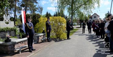 Mogiła, przed którą stoi dwóch mężczyzn w mundurach strazy miejskiej. Przed nimi stoi mężczyzna w garniturze, zwrócony do grupy ludzi w różnym wieku.