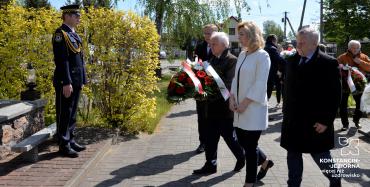 Trzech mężczyzn w czarnych płaszczach i kobieta w białym płaszczy. Jeden z mężczyzn trzyma w dłoniach wieniec z biało-czerwonych kwiatów.