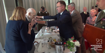Starszy mężczyzna całuje szablę, która podtrzymuje stojący na przeciwko niego nieco młodszy. Są ubrani w garnitury, między nimi jest nakryty do posiłku długi stół.