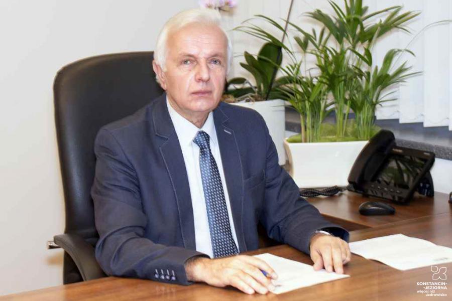 Starszy mężczyzna w biurze, ubrany w garnitur, siedzi przy biurku, na którym leżą dokumenty i stoją telefon oraz kwiaty w doniczce. W ręku trzyma długopis i patrzy się w obiektyw.