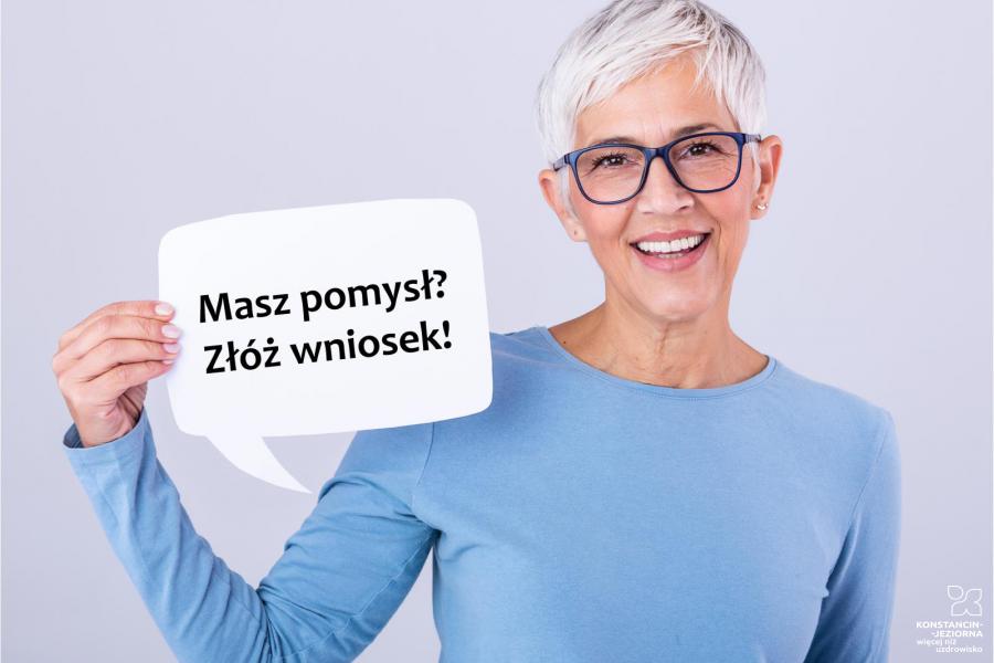 Na zdjęciu uśmiechnięta kobieta – seniorka (krótkie siwe włosy, czarne okulary, ubrana jest w niebieską bluzkę), kobieta trzyma tabliczkę, z napisem: w pierwszym wierszu: Masz pomysł? w drugim wierszu: Złóż wniosek!