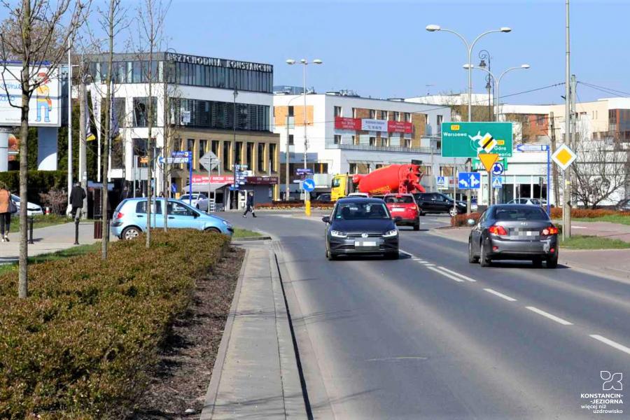 Ulica dwukierunkowa w Konstancinie-Jeziornie. Po ulicy jadą samochody, po chodnikach idą piesi. W dali widać budynki oraz duże banery reklamowe. 