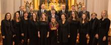 Zdjęcie grupowe Konstancińskiego Chóru Kameralnego. Osiemnaście osób stoi w dwóch rzędach. Ubrani są na czarno. Zdjęcie zrobione w kościele przed ołtarzem. 