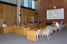 Sala konferencyjna ratusza. Przy trzech prostokątnych stołach, ustawionych w odwróconą literę U, stoi po dłuższych bokach kilka beżowych krzeseł, na niektórych siedzi kilkanaście osób. Na ścianie z czerwonej cegły wisi ekran, na którym wyświetlany jest obraz z sali.