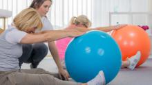 Sala fitness. Dwie kobiety siedzą na podłodze, przed sobą mają duże kolorowe piłki. Wykonują ćwiczenia. Między nimi stoi instruktorka. 