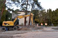 Remont drogi. Widać rozkopaną ziemię, obok stoi żółta koparka. Przy koparce stoi dwóch robotników, którzy ubrani są w pomarańczowe kurtki z odblaskami.